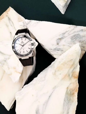 Omega Constellation wristwatch with a black leather strap on white marble fragments against a dark green background in a still life.