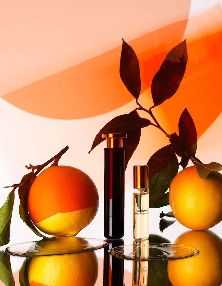 Two perfume bottles and two oranges with green leaves arranged on a reflective surface with a warm orange and white gradient background.