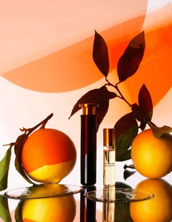 Two perfume bottles and two oranges with green leaves arranged on a reflective surface with a warm orange and white gradient background.