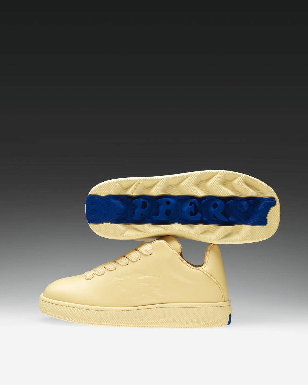Cream colored Burberry sneakers with light tan soles and blue logo on sole against a gradient gray and white background. 