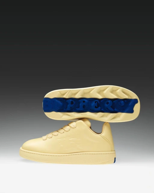 Cream colored Burberry sneakers with light tan soles and blue logo on sole against a gradient gray and white background. 