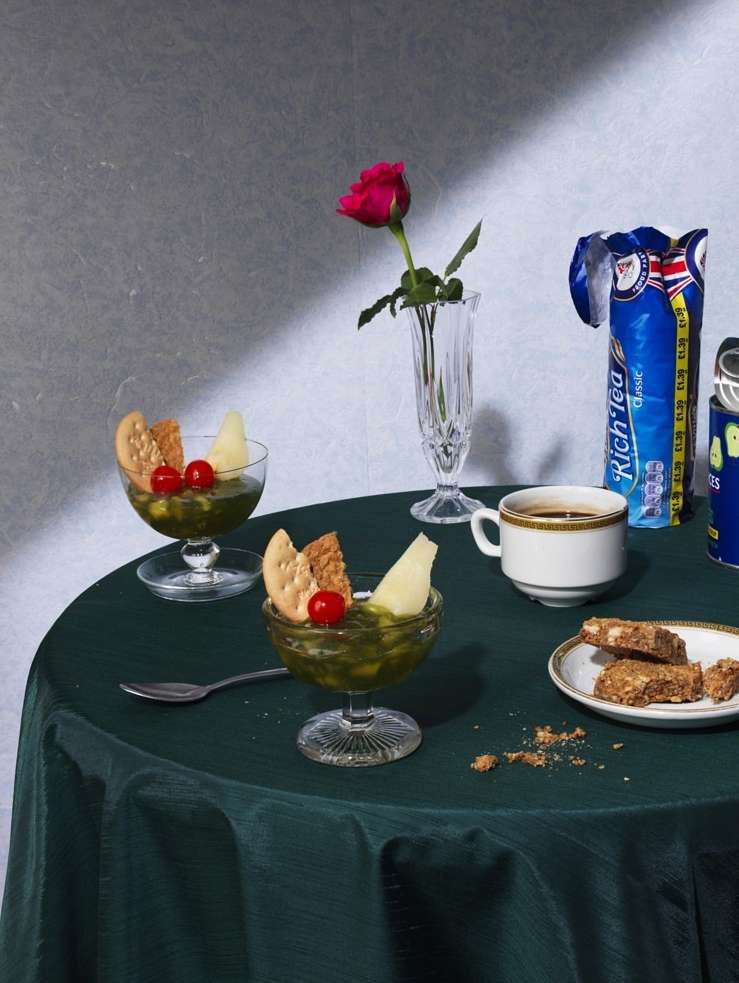 Still life with two dessert glasses of green puree, biscuits, cherries, rose in vase, coffee, and Rich Tea biscuit packaging on a table.