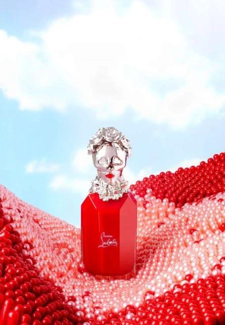 Elegant red perfume bottle with silver skull cap and decorative floral elements sits on a bed of red and white pearl beads.