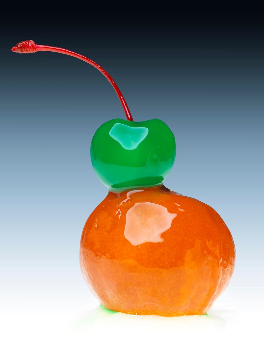 Candied cherry with a green top and red stem against a gradient blue background in a food still life studio photograph.