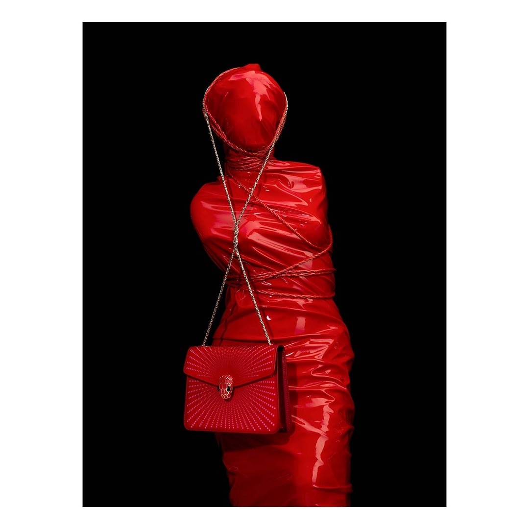 Red-wrapped mannequin torso displays a matching Bulgari purse with a gold chain on a black background in a studio shot.