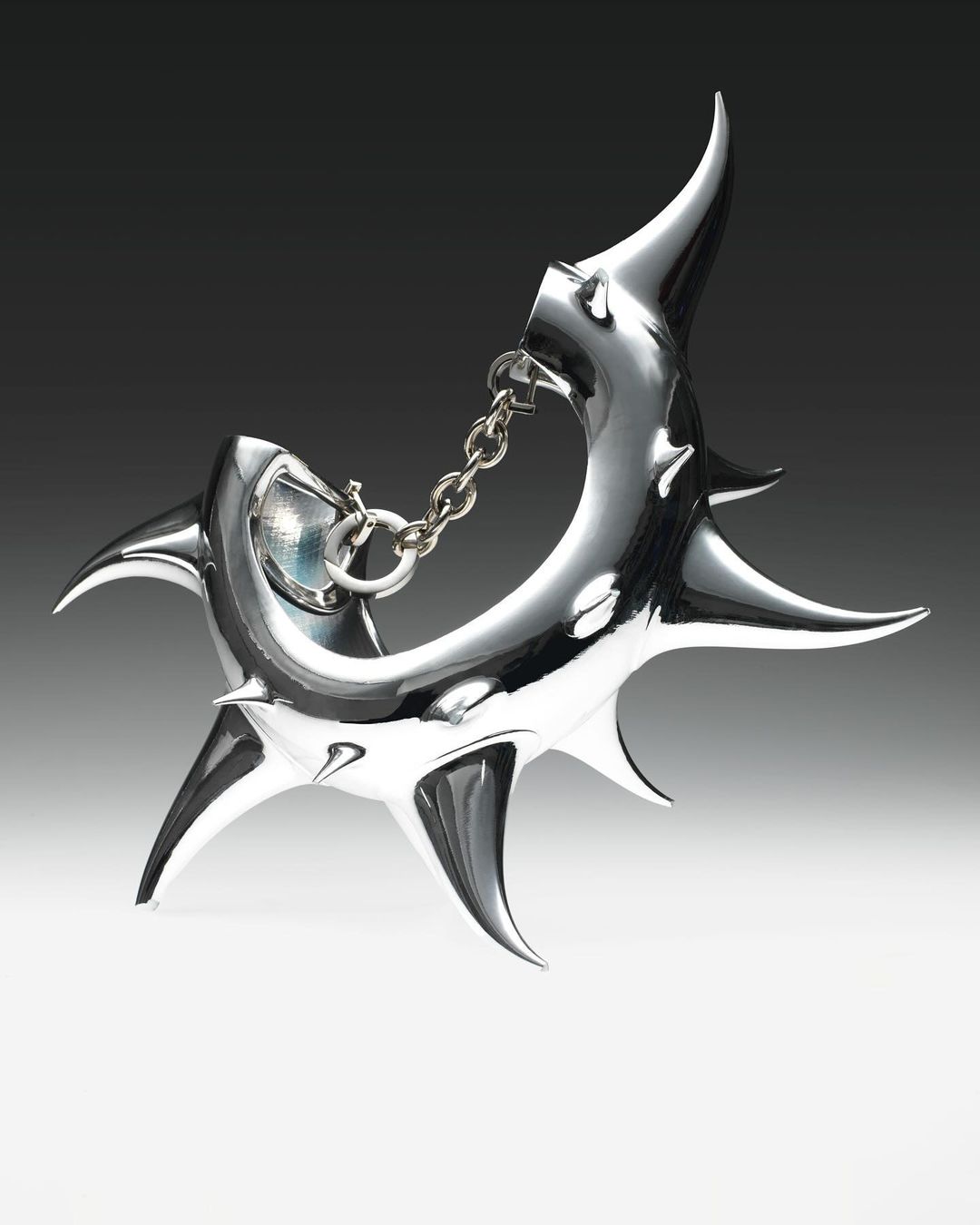 Close-up of a shiny, silver-colored abstract metal sculpture with pointed projections and a chain connection on a gradient background.