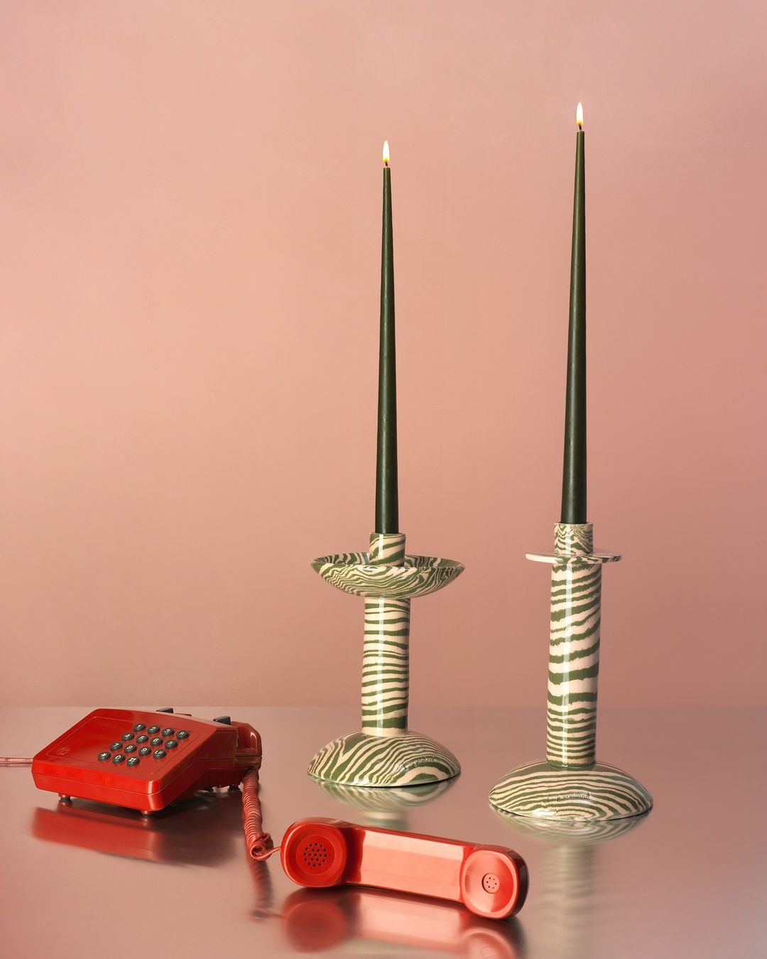 Still life with two black taper candles in patterned ceramic holders and a red rotary telephone on a reflective silver surface.
