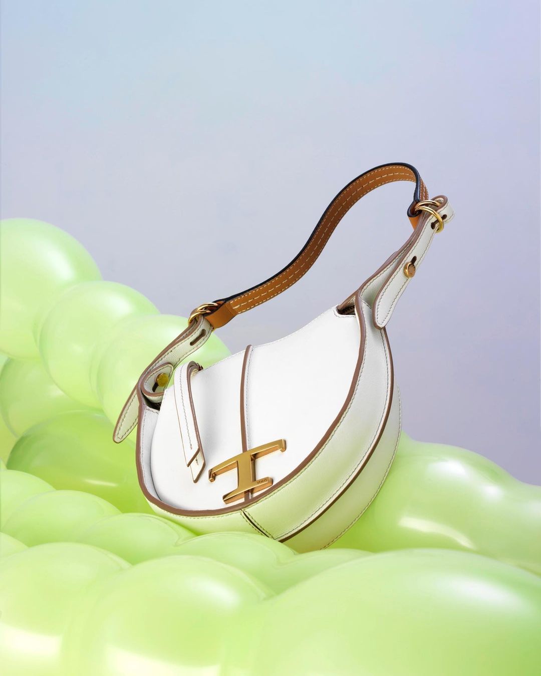 White leather Tod's handbag featuring the brand's logo on a bed of green balloons, presented on a soft gradient studio backdrop.
