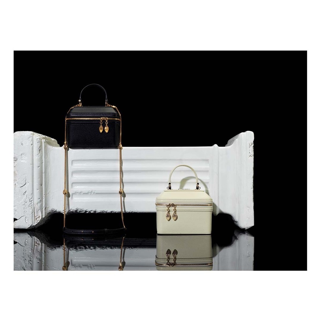 Two structured box purses with gold chain straps on a reflective surface against a black background. One purse is black, the other cream.
