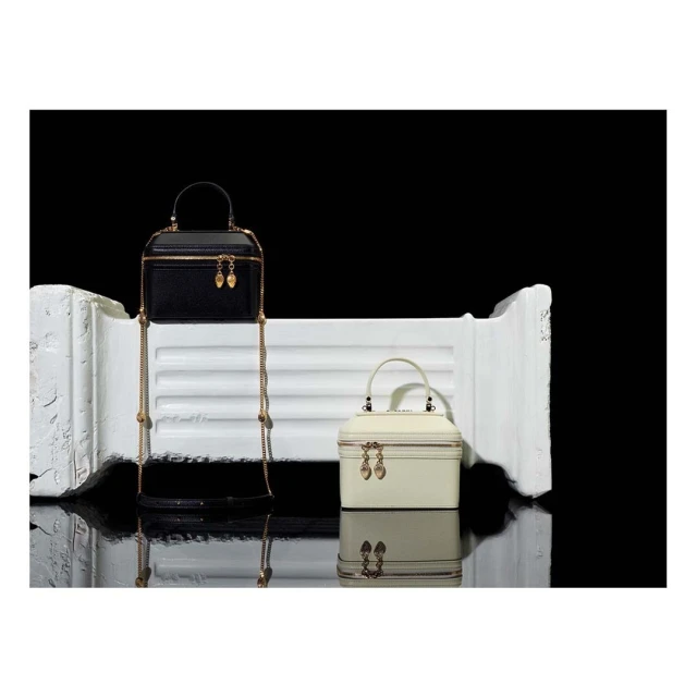 Two structured box purses with gold chain straps on a reflective surface against a black background. One purse is black, the other cream.