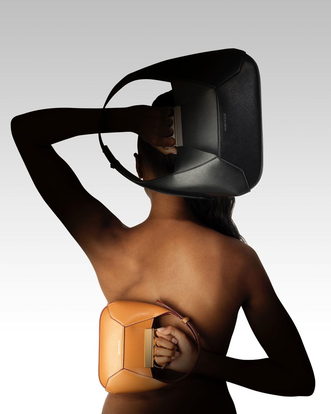 Back view of a topless person holding two leather handbags: a black handbag held over their head, and a tan handbag behind their back.