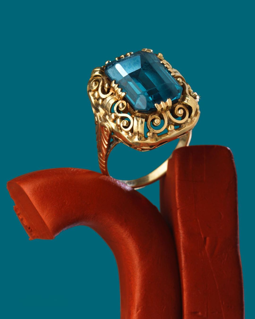 Close-up studio shot of a gold ring with a large rectangular blue gemstone, resting on a red, rounded support against a teal background.