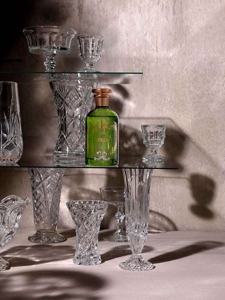 Still life showcases a green perfume bottle surrounded by clear crystal glassware of various sizes on glass shelves against a grey wall.