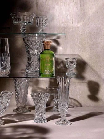 Still life showcases a green perfume bottle surrounded by clear crystal glassware of various sizes on glass shelves against a grey wall.