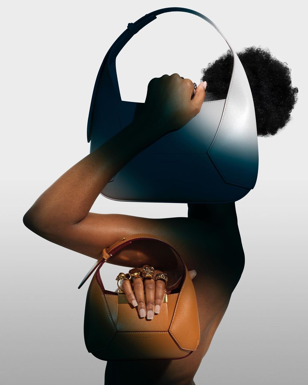 Woman poses with a large blue-grey purse held above her head and a small brown purse in her other hand against a gradient white background.