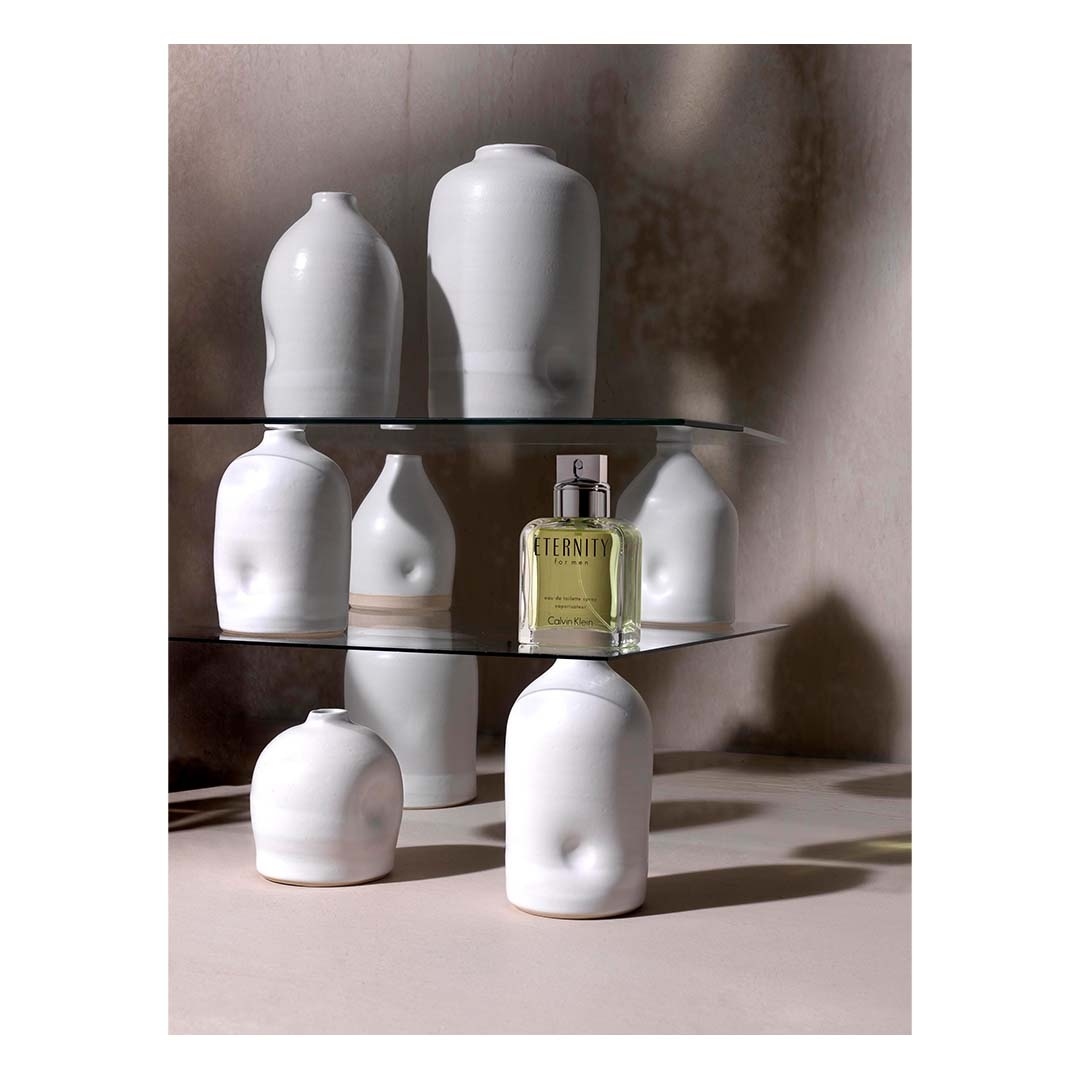 Still life captures a perfume bottle amidst white ceramic vases on glass shelves against a neutral backdrop, creating a minimalist aesthetic.