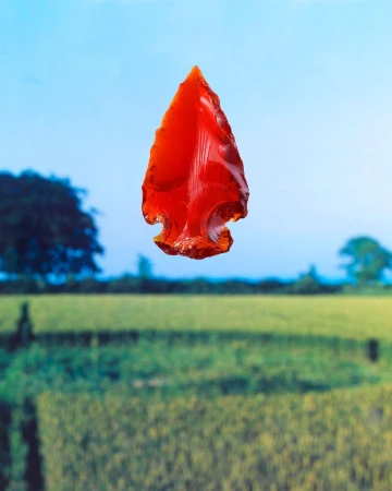 Vibrant red translucent arrowhead suspended against a blurred green field and clear blue sky horizon.
