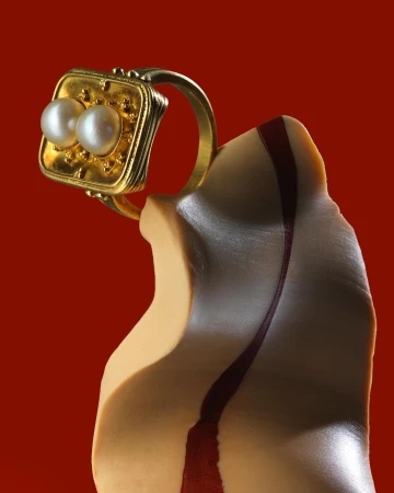 Close up product shot of a gold ring with two pearls resting on an abstract sculptural form against a solid dark red background