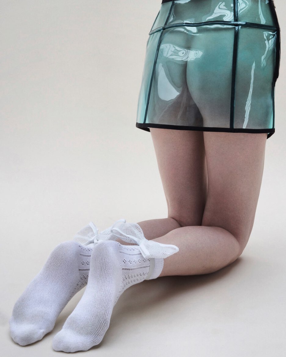 Back view of a woman kneeling wearing a teal, transparent mini skirt with black trim, and white socks with bows, on a plain background.