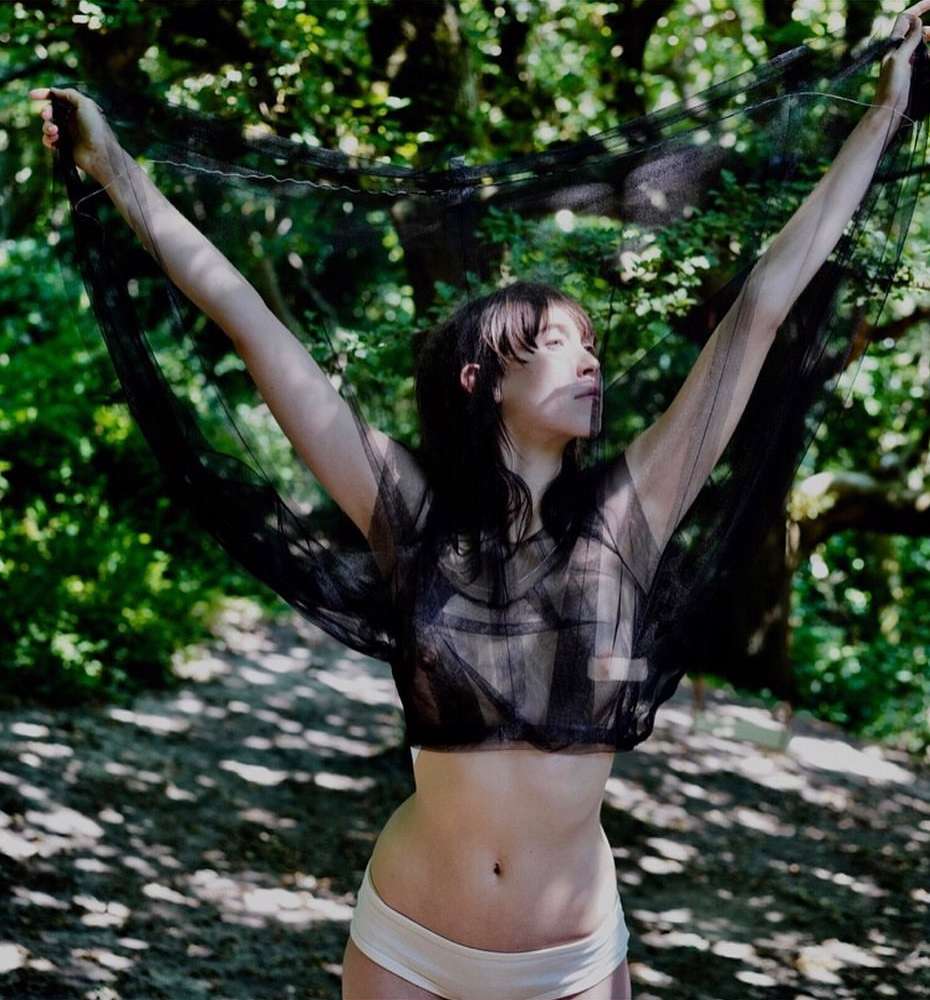 Woman posing with arms raised, wearing a black mesh top and light-colored briefs in a forest setting with dappled sunlight.