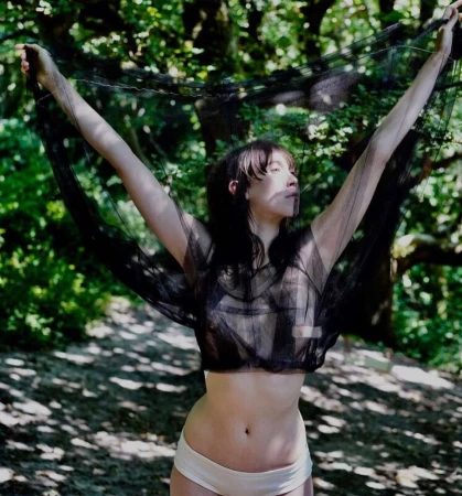 Woman posing with arms raised, wearing a black mesh top and light-colored briefs in a forest setting with dappled sunlight.