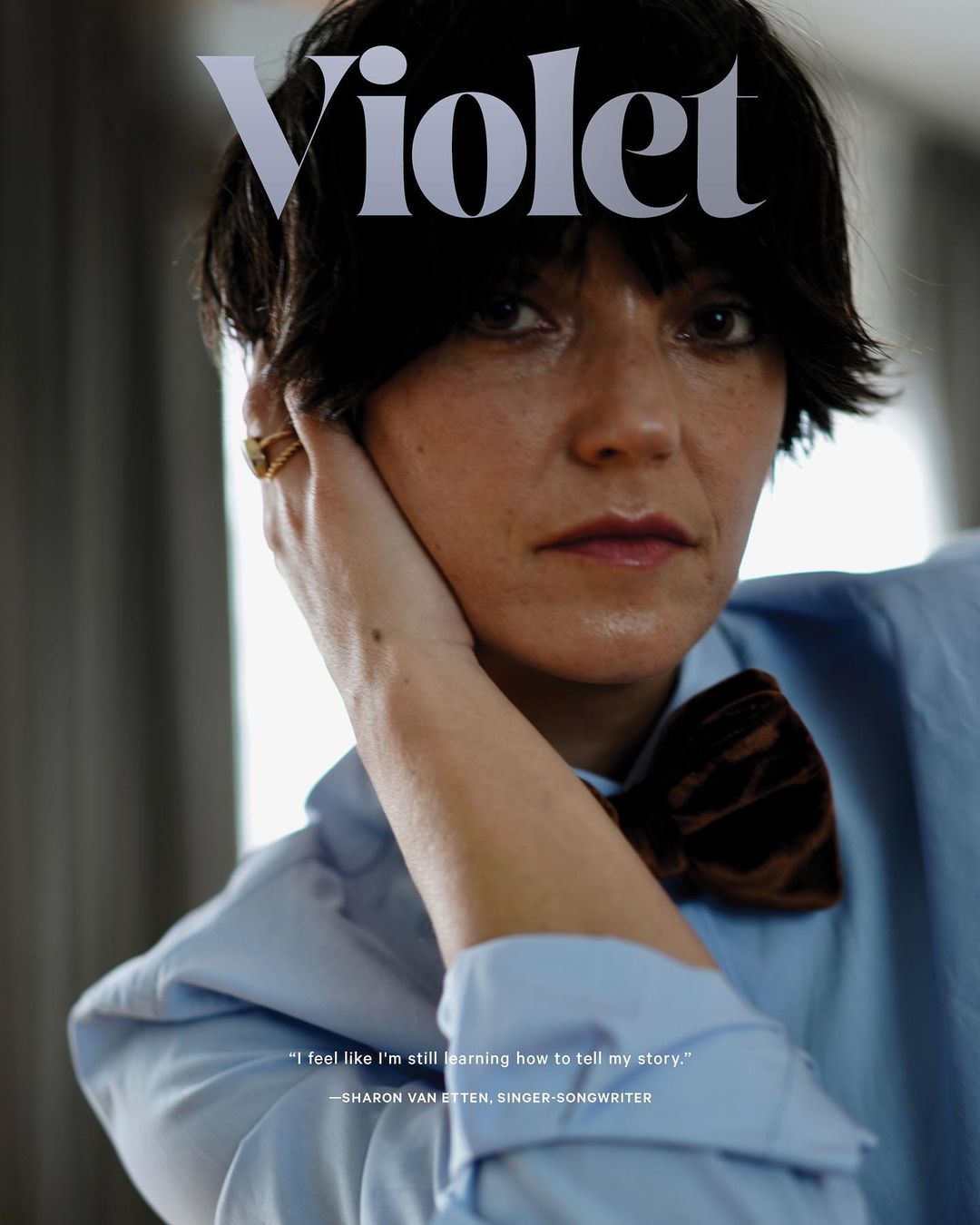 Close-up of a woman with short dark hair, wearing a light blue shirt and brown velvet bow tie. Magazine cover 'Violet' visible above her.