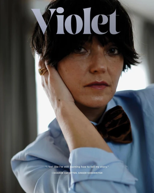 Close-up of a woman with short dark hair, wearing a light blue shirt and brown velvet bow tie. Magazine cover 'Violet' visible above her.