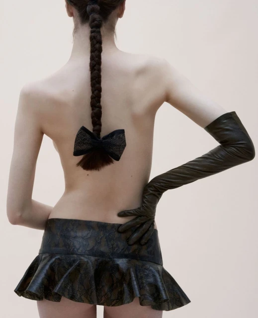 Back view of a slender adult woman with a braided hair and a black bow, wearing a dark leather miniskirt and elbow-length gloves in a studio.