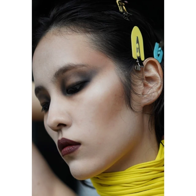 Close-up studio shot of a model with dark eyeshadow and lipstick, hair clips, and a bright yellow rope necklace against a black backdrop.