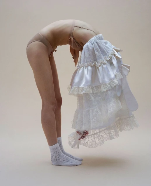 Woman in underwear arches backward, holding ruffled white fabric, creating a visually striking contrast against a neutral backdrop.