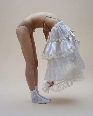 Woman in underwear arches backward, holding ruffled white fabric, creating a visually striking contrast against a neutral backdrop.