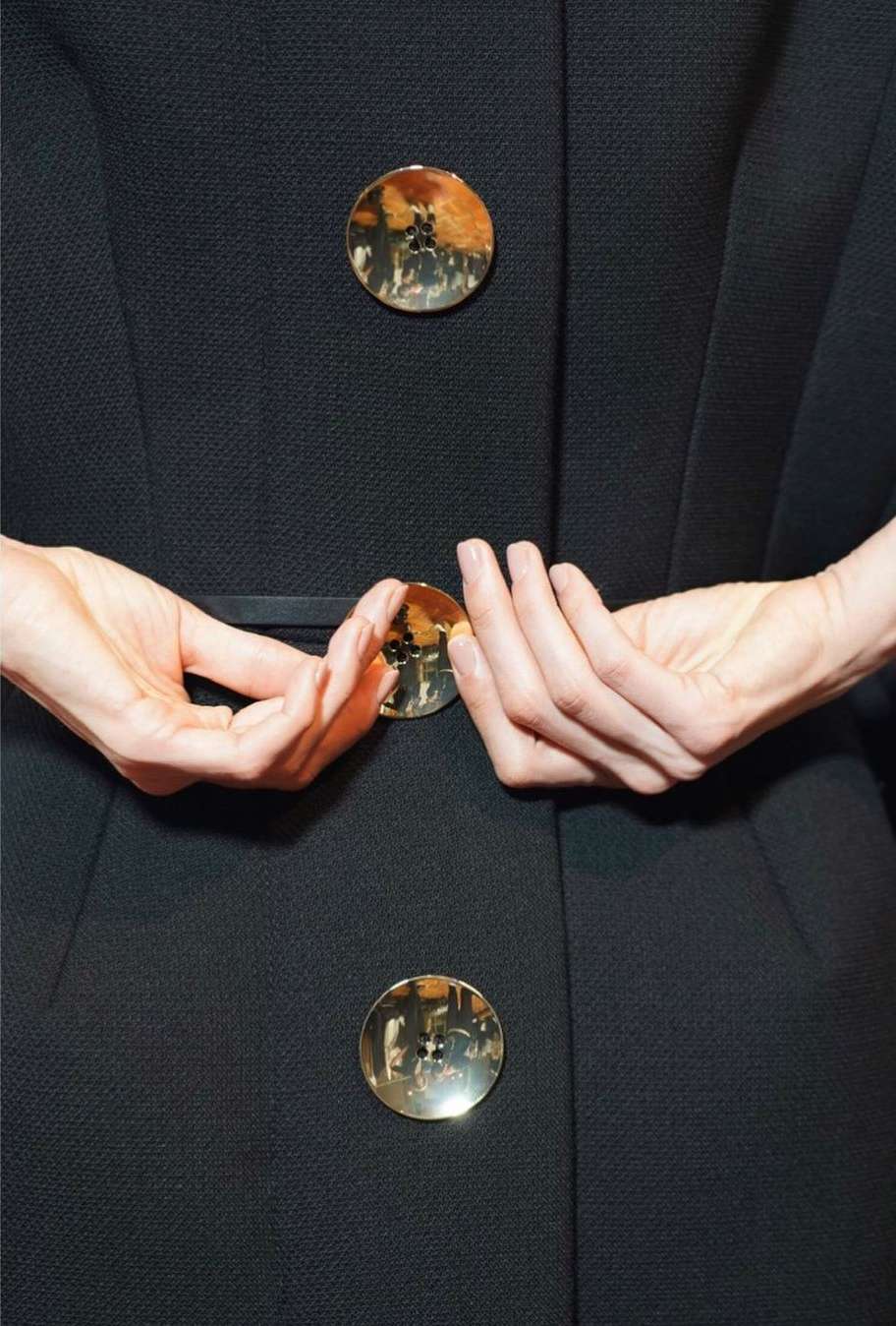 Close-up of a woman's hands holding a black coat with round gold buttons, showcasing fashion and design details indoors.