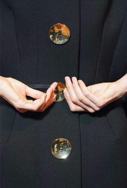 Close-up of a woman's hands holding a black coat with round gold buttons, showcasing fashion and design details indoors.