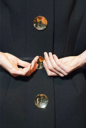 Close-up of a woman's hands holding a black coat with round gold buttons, showcasing fashion and design details indoors.