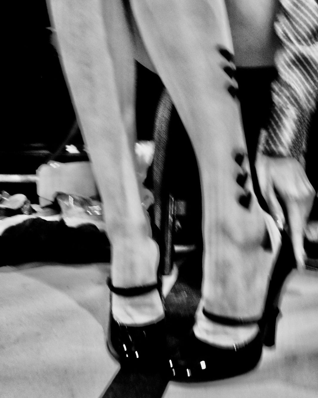 Close-up black and white shot of a woman's legs, high heels, hosiery with heart detail, and part of striped jacket is visible.