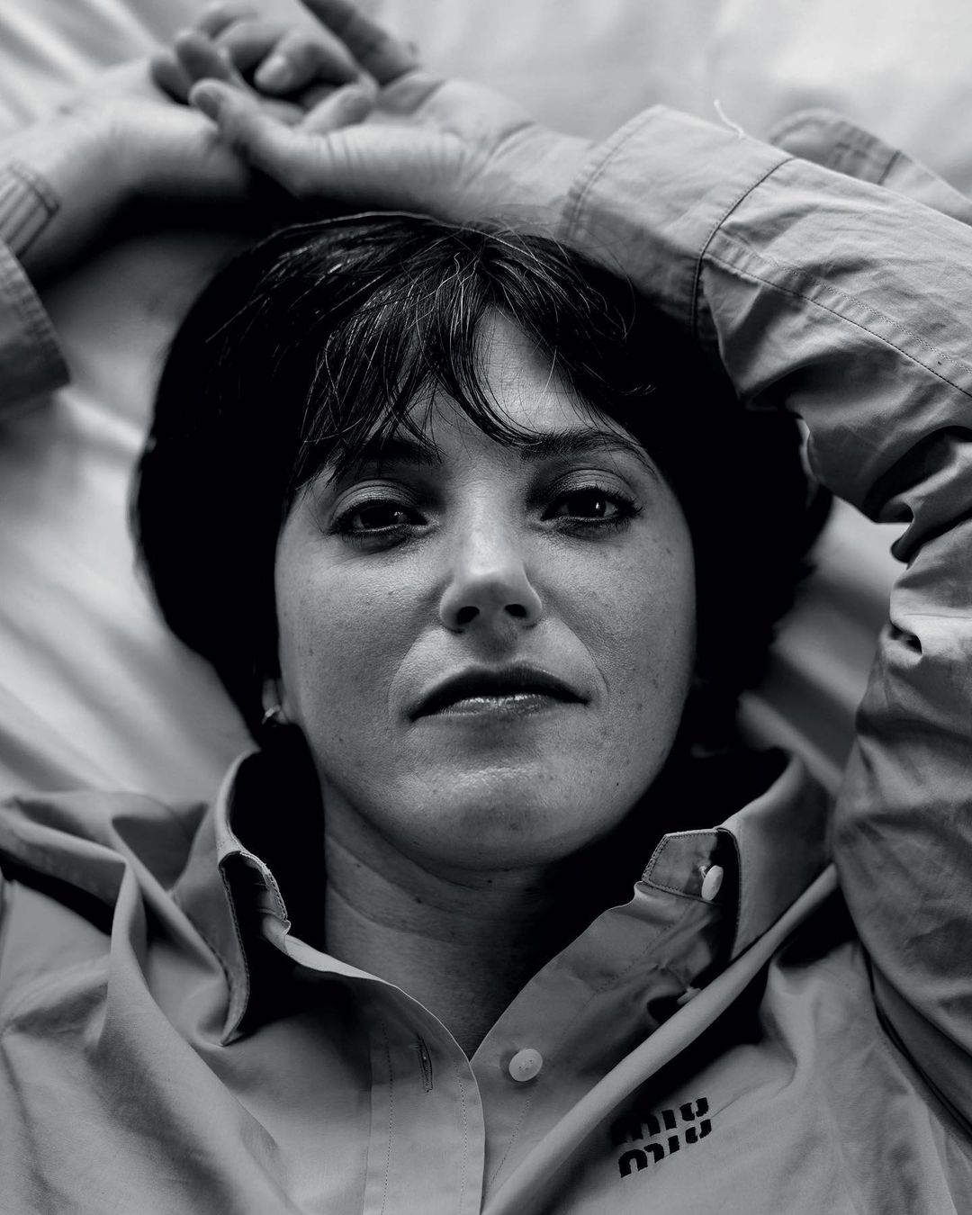 Close-up black and white portrait of a woman with dark hair and bangs lying on a bed wearing a button-down shirt with a logo.