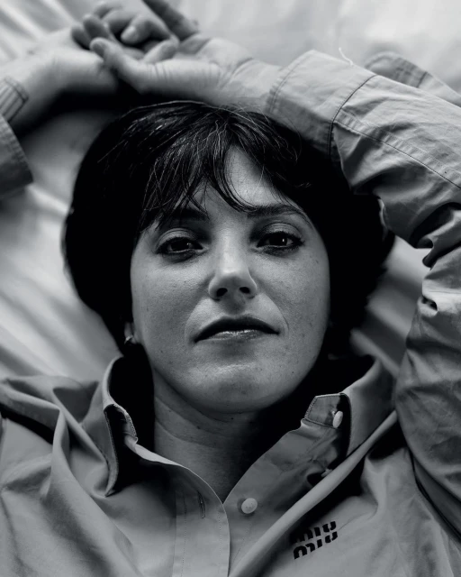 Close-up black and white portrait of a woman with dark hair and bangs lying on a bed wearing a button-down shirt with a logo.