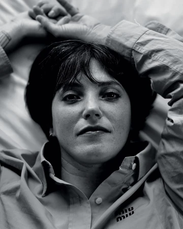 Close-up black and white portrait of a woman with dark hair and bangs lying on a bed wearing a button-down shirt with a logo.