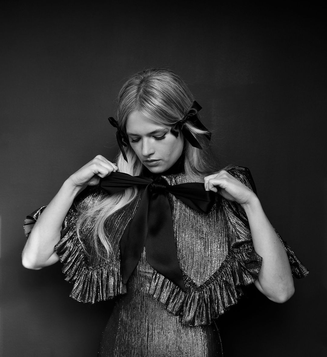 Black and white portrait of a blonde woman tying a large bow at her chest while wearing a shimmering ruffled dress with bows in her hair.