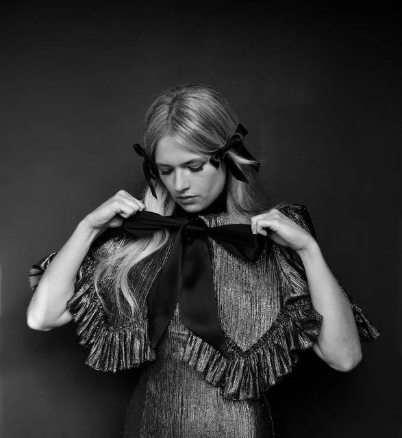 Black and white portrait of a blonde woman tying a large bow at her chest while wearing a shimmering ruffled dress with bows in her hair.