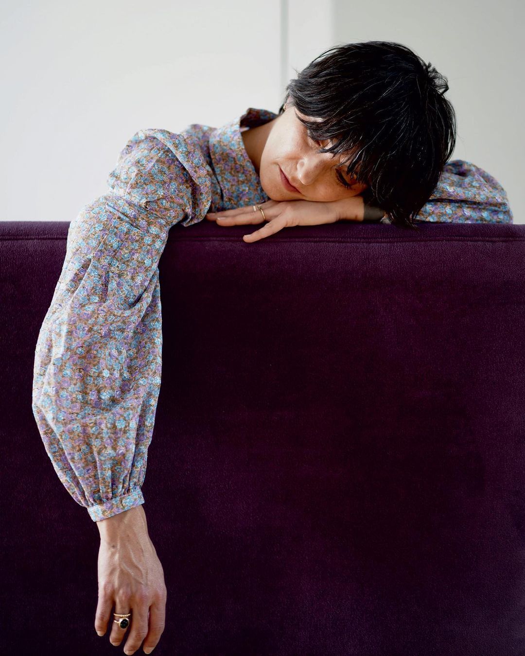 A brunette woman with her head resting on a purple couch, wearing a floral top, in a medium shot with indoor natural lighting.