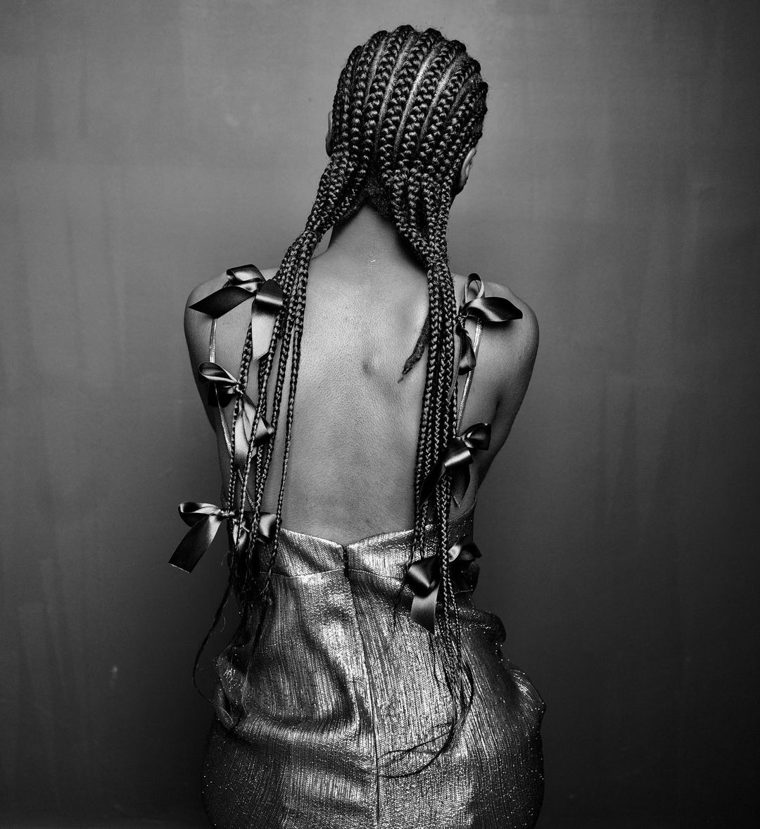 Back view of a woman with braided hair adorned with satin bows, wearing a shimmering dress against a neutral background in black and white.
