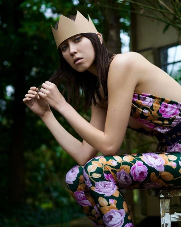 Young woman with cardboard crown poses in a floral top and pants against a blurred greenery background in a medium shot.