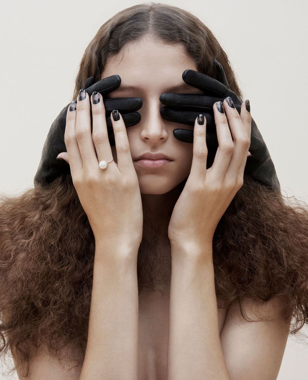Model with long brown curly hair covering her eyes with black leather gloves while wearing a pearl ring in a portrait by Lynette Garland.