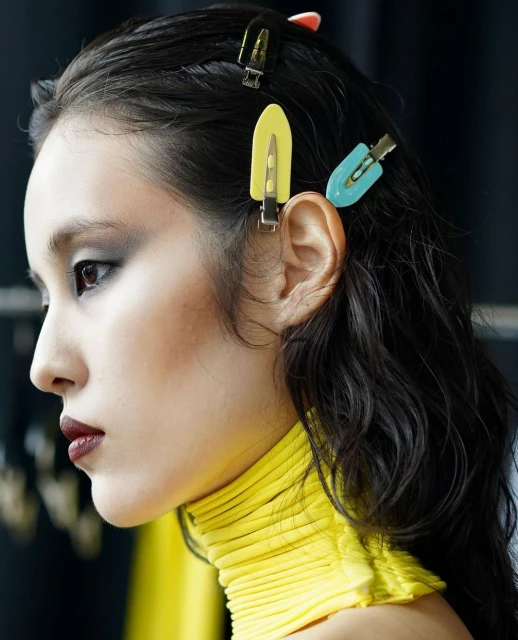 Profile of an Asian woman with dark hair and colorful hair clips, wearing a yellow ribbed turtleneck shirt with a dark background.