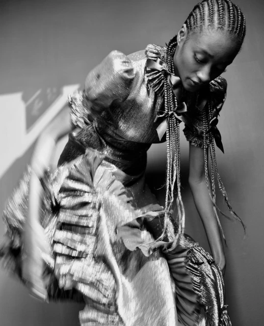 Black and white fashion portrait of a woman in a ruffled, shimmering dress with braided pigtails, posing indoors in a studio with soft lighting.