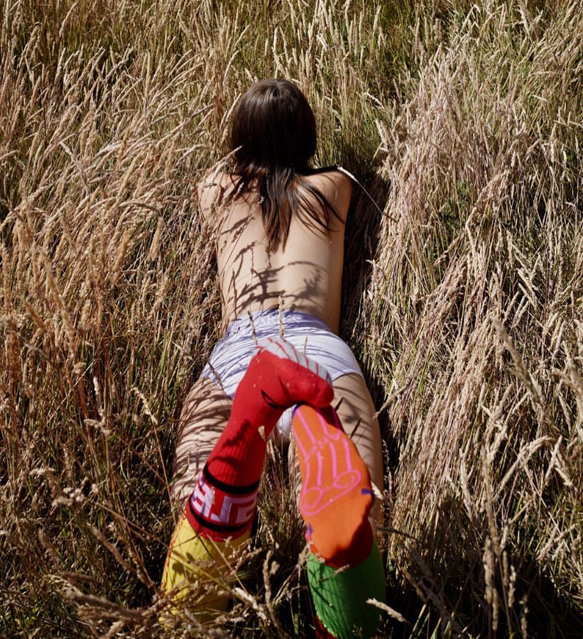Woman lies in golden grass wearing socks. The outdoor shot features natural light and a focus on texture and color contrast.