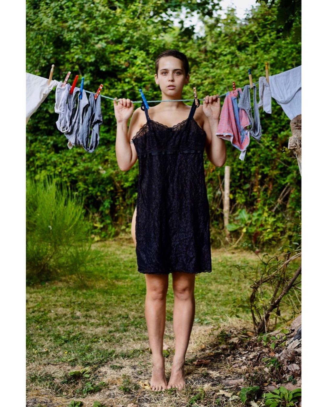 Woman standing outdoors with clothing hanging on a clothesline, featuring black lace dress, greenery background, and natural light.