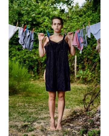 Woman standing outdoors with clothing hanging on a clothesline, featuring black lace dress, greenery background, and natural light.