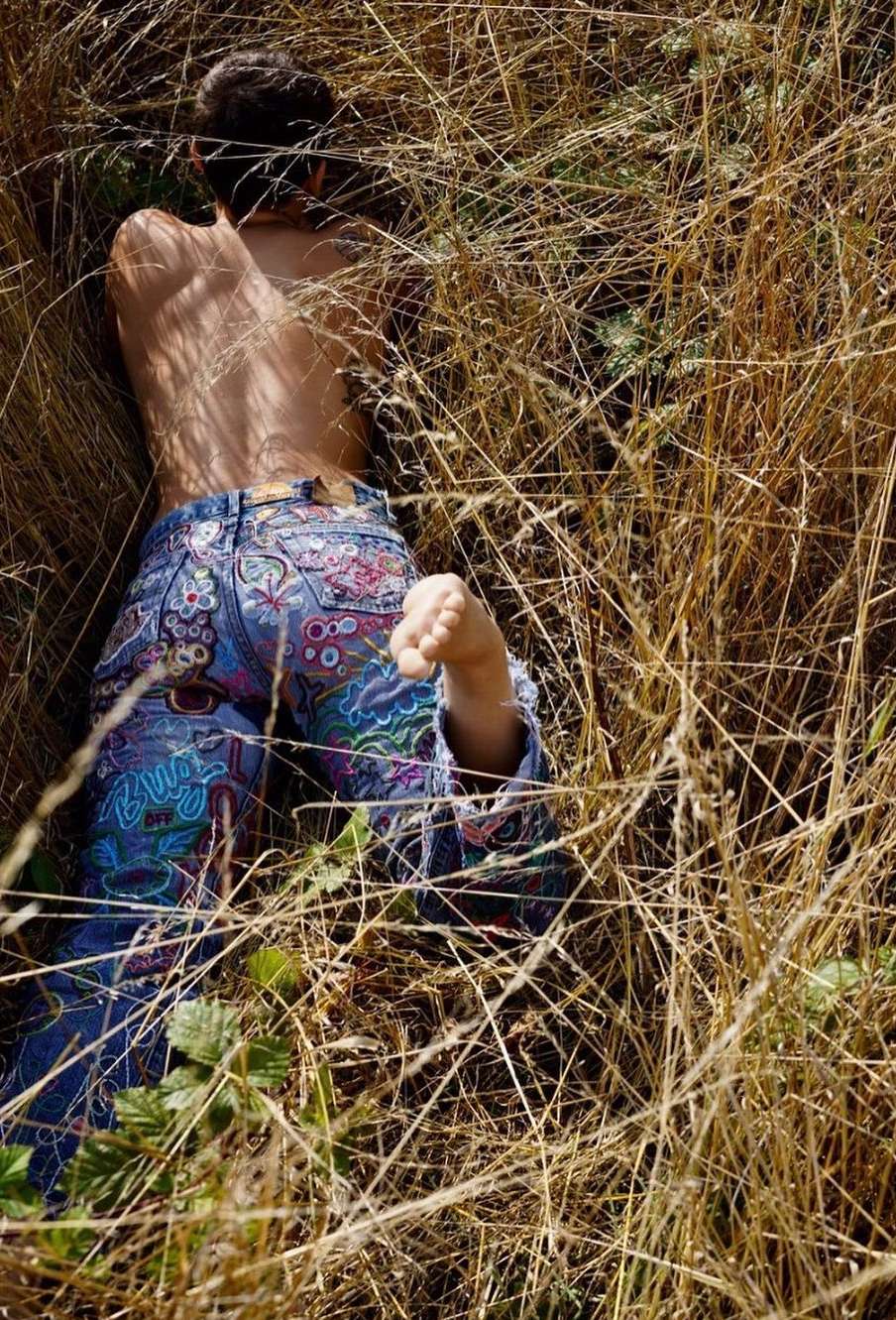 A person lies in dry grasses, showcasing patterned jeans and partial back, in an outdoor nature setting under bright sunlight.